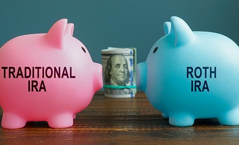 Two Piggy Banks Facing Each Other With Rolled Up Dollar Bills Between Them. One Bank Is Pink And Says Traditional Ira And One Is Blue And Says Roth Ira.