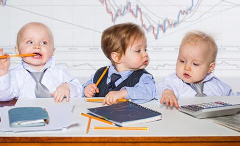 Three young children wearing shirts and ties sitting at a desk holding pencils with notebooks in front of them