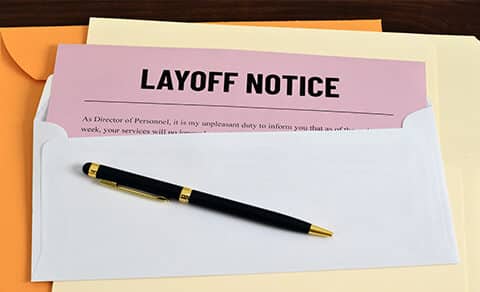 envelope with a pink slip sticking out that says LAYOFF NOTICE, back pen on it. Manila folder underneath envelope
