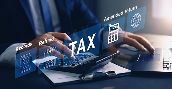 Man in a blue suit working on a laptop and calculator with 5 digitally created screens floating in fron of hm with the words: Records, Refund, Tax, Amended return