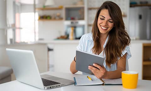 A woman with brown hair and a white top sittind at a desk in her homw ith a laptop and a yellow coffee mug.