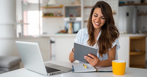 A woman with brown hair and a white top sittind at a desk in her homw ith a laptop and a yellow coffee mug.