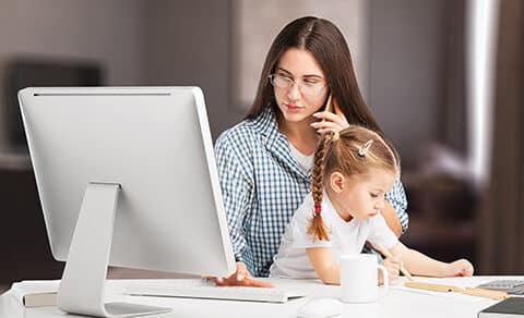 Woman with long brown hair and a blue and white stripe shirt, child wearing a white top and her hair in a ponytail sits on her lap, working on her laprop in her home.