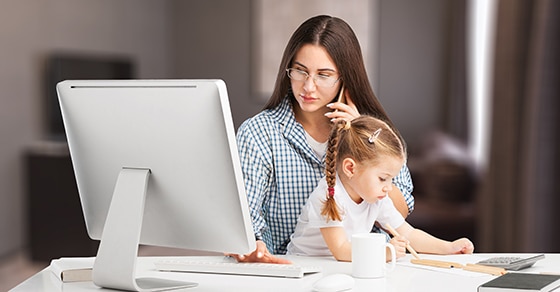 Woman with long brown hair and a blue and white stripe shirt, child wearing a white top and her hair in a ponytail sits on her lap, working on her laprop in her home.