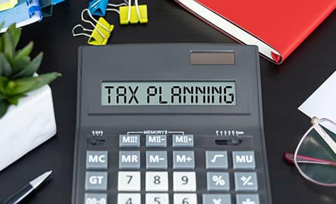 Black desktop, calculaor on it with a read out of TAX PLANNING, eyeglasses, note pads., small pant in a white pot.