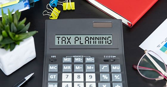 Black desktop, calculaor on it with a read out of TAX PLANNING, eyeglasses, note pads., small pant in a white pot.