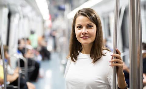 A woman, standing, on a subway, holding a pole, wearing a white shirt, looking confident, passengers in the background.