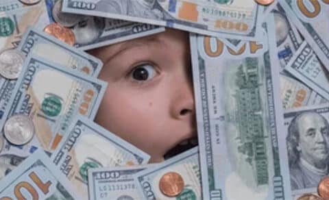The, picture, shows, a, surprised, or, wide-eyed, child's, face, surrounded, by, scattered, U.S., hundred-dollar, bills, quarters, dimes, nickels, and, pennies. The, child’s, expression, suggests, amazement, shock, or, excitement, as, they, peek, through, the, pile, of, money, and, coins.
