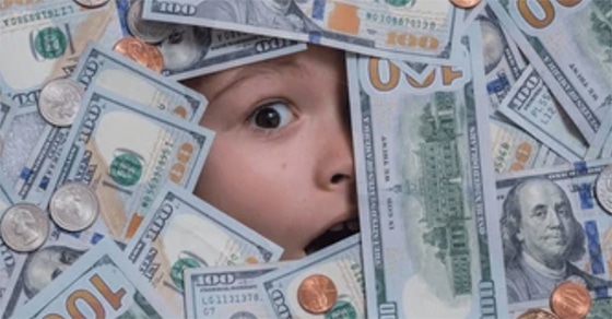 The, picture, shows, a, surprised, or, wide-eyed, child's, face, surrounded, by, scattered, U.S., hundred-dollar, bills, quarters, dimes, nickels, and, pennies. The, child’s, expression, suggests, amazement, shock, or, excitement, as, they, peek, through, the, pile, of, money, and, coins.