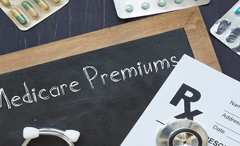 The, image, shows, a, chalkboard, with, "Medicare, Premiums", written, on, it, surrounded, by, pills, a, prescription, pad, and, a, stethoscope, highlighting, healthcare, costs.