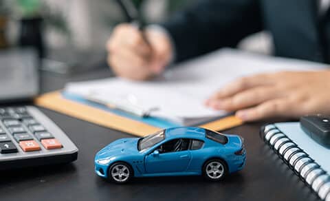 ChatGPT said: A, desk, with, a, blue, toy, car, calculator, car, keys, and, a, person, in, the, background, writing, on, paperwork, suggesting, car, financing, or, purchasing.