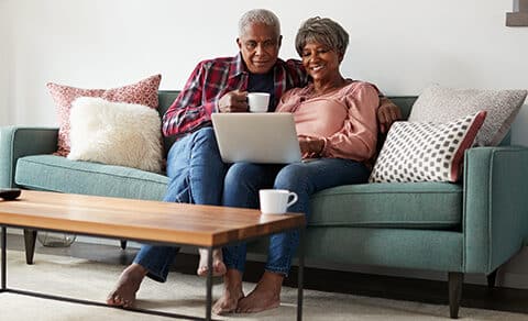 An, older, couple, sits, on, a, sofa, smiling, and, enjoying, coffee, while, looking, at, a, laptop, together.
