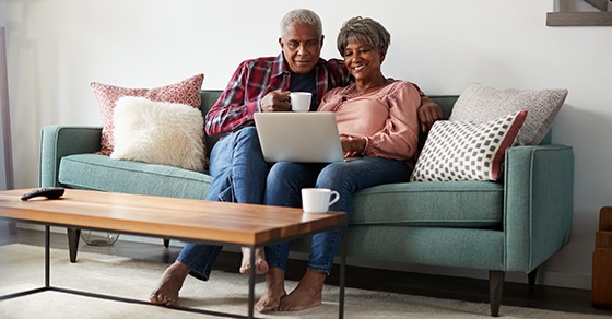 An, older, couple, sits, on, a, sofa, smiling, and, enjoying, coffee, while, looking, at, a, laptop, together.