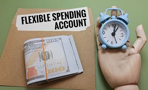 A, folder, labeled, “Flexible, Spending, Account”, sits, on, a, table, with, a, stack, of, cash, on, top,, while, a, wooden, hand, holds, a, small, blue, alarm, clock,, symbolizing, the, urgency, to, use, FSA, funds, before, time, runs, out,.